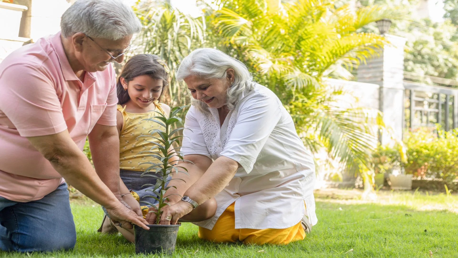 Indian grandparents and granddaughter gardening together outdoors, planting flowers in sunny garden - representing multi-generational family connections and the joyful, engaged lifestyle ElderWorld helps families maintain