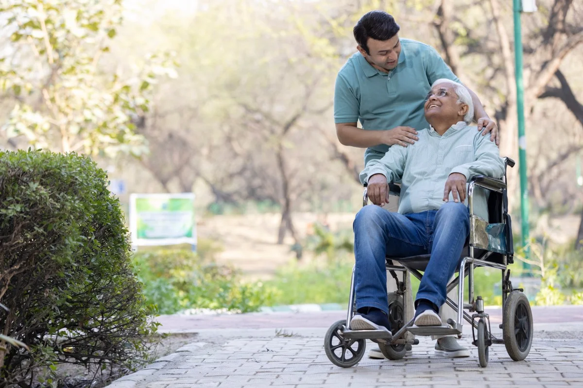 Ongoing care - Caregiver providing compassionate outdoor assistance to elderly person in wheelchair at park, representing regular care activities