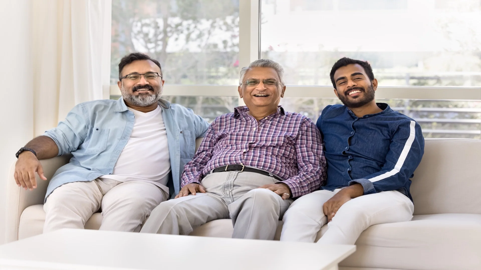 Happy three-generation Indian family portrait with grandfather, father, and grandson smiling together on couch at home, representing satisfied families who trust ElderWorld for senior care in Bangalore